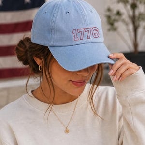 May include: Light blue baseball cap with the number "1776" embroidered in red. The cap is made of a soft material and has a curved brim. The background includes an American flag.