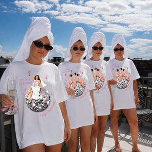 May include: A white t-shirt featuring a graphic of a person in a white dress sitting on a disco ball, with the text "EST 2026 MIAMI FLORIDA" and "Emily's Bachelorette". The graphic includes pink star accents and a cocktail glass illustration.