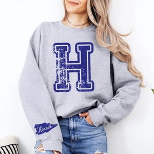 Op de afbeelding: Een lichtgrijs sweatshirt met lange mouwen en een grote, verweerde donkerblauwe letter H in het midden op de borst. Op de linkermouw staat een kleine donkerblauwe wimpelafbeelding met de naam Hannah in een wit cursief lettertype.