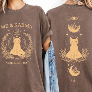 Op de afbeelding: Bruin t-shirt met een ontwerp voor en achter. De voorkant heeft de tekst "ME & KARMA VIBE LIKE THAT" met een kattenillustratie. De achterkant heeft een kattenillustratie, ogen, maan- en sterontwerpen.