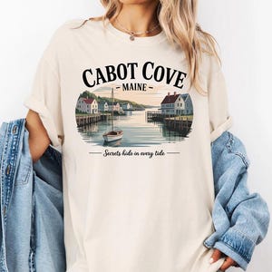 A woman wearing a white t-shirt with a graphic design depicting Cabot Cove, Maine, featuring a boat on a lake and houses along the shoreline.