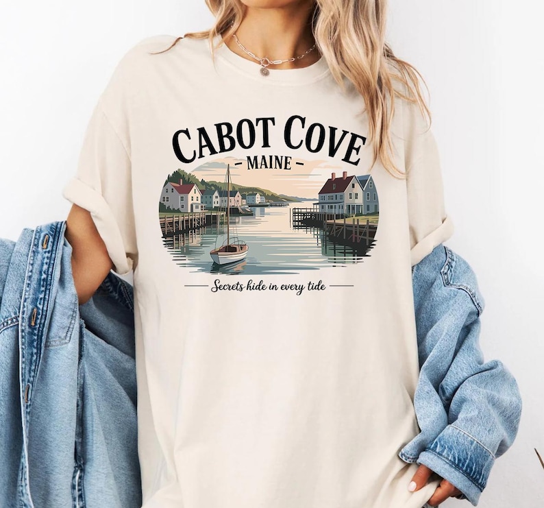 A woman wearing a white t-shirt with a graphic design depicting Cabot Cove, Maine, featuring a boat on a lake and houses along the shoreline.