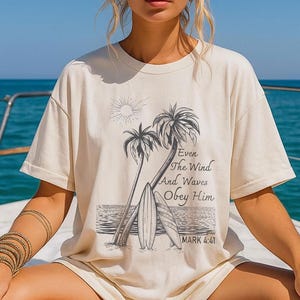 A woman wearing a white t-shirt with a graphic design of palm trees and a beach scene, along with a Bible verse from the book of Mark.