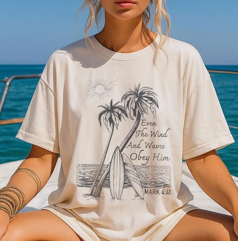 A woman wearing a white t-shirt with a graphic design of palm trees and a beach scene, along with a Bible verse from the book of Mark.