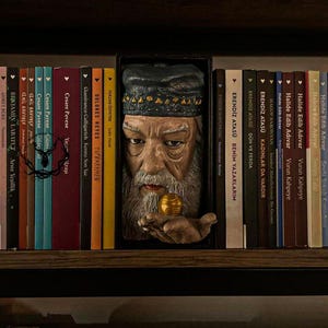 May include: A wooden bookshelf displaying a carved sculpture of an elderly man with a beard and hat, holding a golden orb. The sculpture is positioned between a row of colourful books with Turkish titles.