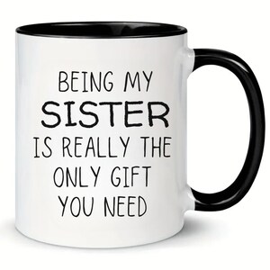 Puede incluir: Taza de café de cerámica blanca con borde y asa negros. La taza tiene texto negro que dice "Being my sister is really the only gift you need."