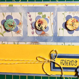 May include: A handmade greeting card featuring a bright yellow base with a blue panel displaying three paper flowers. Each flower has a circular center with bird and floral patterns. A yellow and white twine bow sits near a label reading FRIENDS.