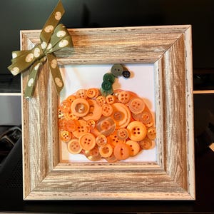 May include: A square frame with a wood-grain finish holds a pumpkin-shaped mosaic made of various orange buttons. A green button stem sits atop the pumpkin, and a green ribbon with a white pumpkin pattern is tied to the top left corner of the frame.