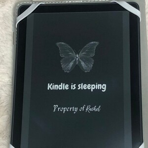 May include: A black Kindle e-reader with a white butterfly graphic on the screen. The text "Kindle is sleeping" and "Property of Rachel" is displayed on the screen.