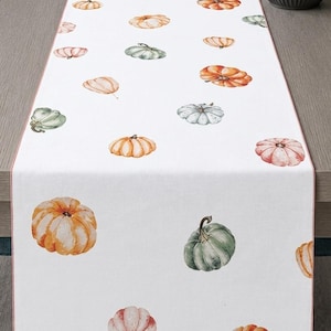May include: A white table runner featuring a repeating pattern of watercolour-style pumpkins in various shades of orange, sage green, and muted pink. The rectangular runner has a thin, solid-coloured border along its long edges and a smooth finish.
