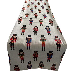 May include: A rectangular table runner with a repeating pattern of toy nutcracker soldiers in red, blue, black, and gold uniforms on a light gray background. The runner is made of fabric and is suitable for holiday decor.