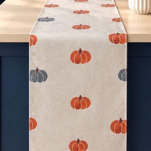 May include: A rectangular table runner made of light beige fabric featuring a repeating pattern of printed pumpkins. The pumpkins are coloured in shades of burnt orange and muted slate blue, arranged in a grid-like layout across the entire surface.