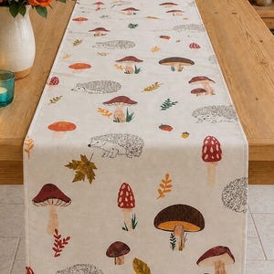 May include: A rectangular table runner featuring a repeating pattern of hedgehogs, various mushrooms in shades of red, brown, and orange, and scattered autumn leaves on a light cream-coloured fabric background with finished stitched edges.