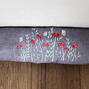 May include: A long, rectangular door draft excluder in a dark grey fabric. The excluder is embroidered with a floral design featuring red flowers and white accents. It rests on a wooden floor.