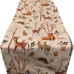 May include: A white table runner with a woodland animal print. The print features deer, foxes, rabbits, birds, and trees in shades of brown, orange, and green.