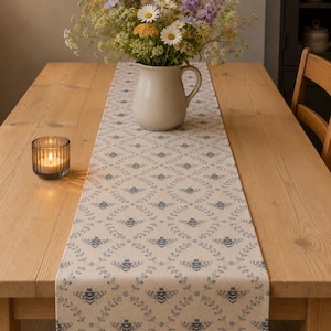 Peut inclure: Chemin de table beige avec un motif répété d'abeilles et de feuilles bleues. Le chemin de table est posé sur une table en bois clair, avec un vase de fleurs et une bougie allumée dans un photophore. Les fleurs sont blanches, jaunes et violettes.