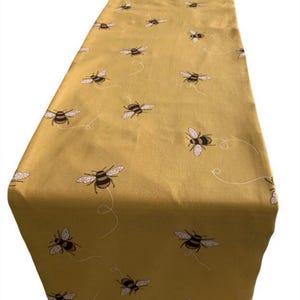 May include: A yellow table runner with a repeating pattern of black and brown bees. The bees are flying and have a white outline.