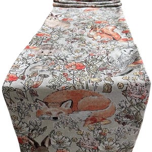 May include: A grey linen table runner with a woodland animal print. The runner features a sleeping fox, a rabbit, a squirrel, a hedgehog, mushrooms, and flowers in shades of orange, red, and green.