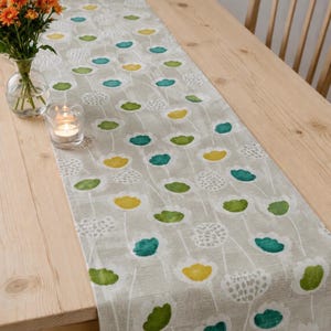 May include: A floral table runner with a light gray background and a repeating pattern of white, yellow, green, and teal flowers. The runner is on a light wood table, with a vase of orange flowers and a lit candle in the background.