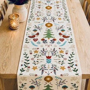 May include: A long rectangular table runner featuring a repeating pattern of green Christmas trees, gold stars, red hearts, blue ornaments, and stylized floral foliage on a cream-colored fabric background, centered on a light wood dining table.