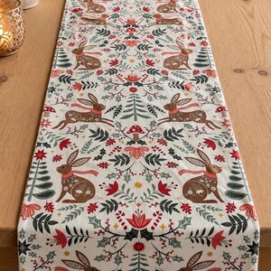 May include: A rectangular table runner featuring a repeating pattern of brown rabbits wearing red scarves, interspersed with red mushrooms, green pine trees, holly leaves, and small red berries on a cream-coloured fabric background.
