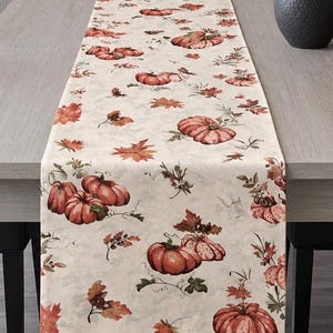 May include: A rectangular table runner featuring a cream-coloured background with a repeating pattern of orange pumpkins, brown autumn leaves, and green foliage. The fabric displays various sizes of pumpkins and scattered fall-themed botanical elements.