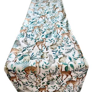 May include: White fabric table runner with a woodland animal pattern. The design includes brown deer, grey rabbits, and owls, interspersed with green foliage. Ideal for a dining table or decorative display.