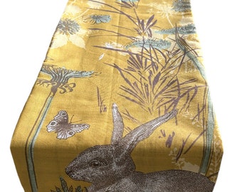 May include: A yellow table runner with a brown rabbit, green leaves, and a butterfly design.