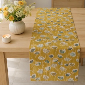 May include: A rectangular table runner featuring a mustard yellow background with a repeating pattern of stylized white and sage green flowers on thin stems. The fabric runner is laid flat across a light-toned wooden table surface.