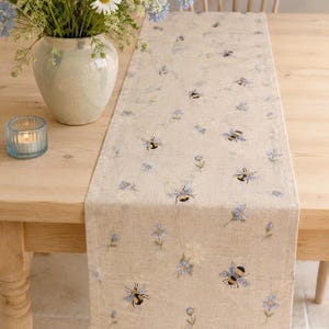 May include: A rectangular table runner made of light beige fabric featuring a repeating pattern of small blue flowers and bumblebees. The runner is laid flat across a wooden table, showcasing the detailed embroidery of the insects and floral motifs.