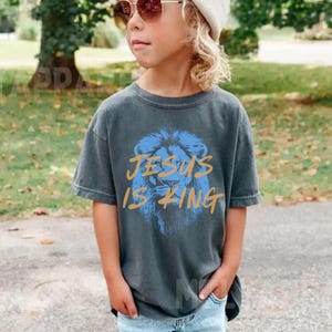 以下が含まれることがあります： 青いライオンのグラフィックと金色の文字「JESUS IS KING」がプリントされたグレーのTシャツを着た子供。 子供はまた、スマイリーフェイスのクリーム色のビーニーと水色のジーンズを着用しています。