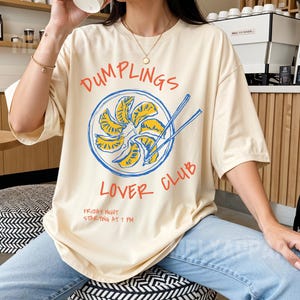 Op de afbeelding: Ivoor T-shirt met een afbeelding van "Dumplings Lover Club". Het ontwerp toont een bord met dumplings en eetstokjes. De tekst "Friday Night Starting at 7 PM" staat ook op het shirt. Het model draagt een lichtblauwe spijkerbroek.