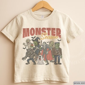 May include: A natural heather short-sleeve t-shirt featuring a graphic print with the text "MONSTER Season" above illustrations of a mummy, witch, vampire, ghost, zombie, werewolf, and Frankenstein's monster, all rendered in a vintage style.