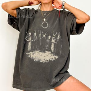May include: Dark gray t-shirt with a graphic print of figures with horns and a sun design. The text "MELY APPAREL" is visible on the shirt. The word "PEPPER" is in a white oval at the bottom.