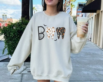 May include: A sand-colored crewneck sweatshirt featuring a graphic print on the chest. The design displays the word BOO, where the letter O is replaced by two teeth illustrations filled with small orange and black Halloween-themed icons.