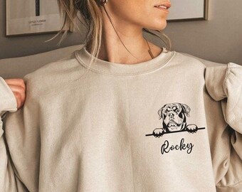 May include: A beige sweatshirt with a black and white illustration of a dog peeking over the top. The text "Rocky" is printed below the dog.