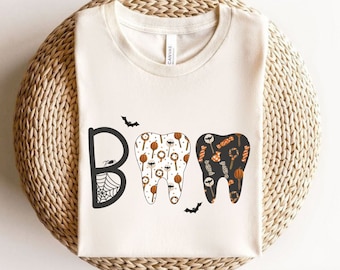 Puede incluir: Camiseta color crema con un diseño temático de Halloween. La palabra "BOO" está escrita con una telaraña y dos dientes decorados con caramelos y calabazas. Murciélagos están dispersos alrededor del diseño.