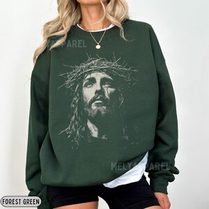May include: Forest green sweatshirt featuring a detailed graphic of Jesus wearing a crown of thorns. The image is a grayscale portrait. The sweatshirt has a crew neck and long sleeves. The text "MELY APPAREL" is visible on the garment.