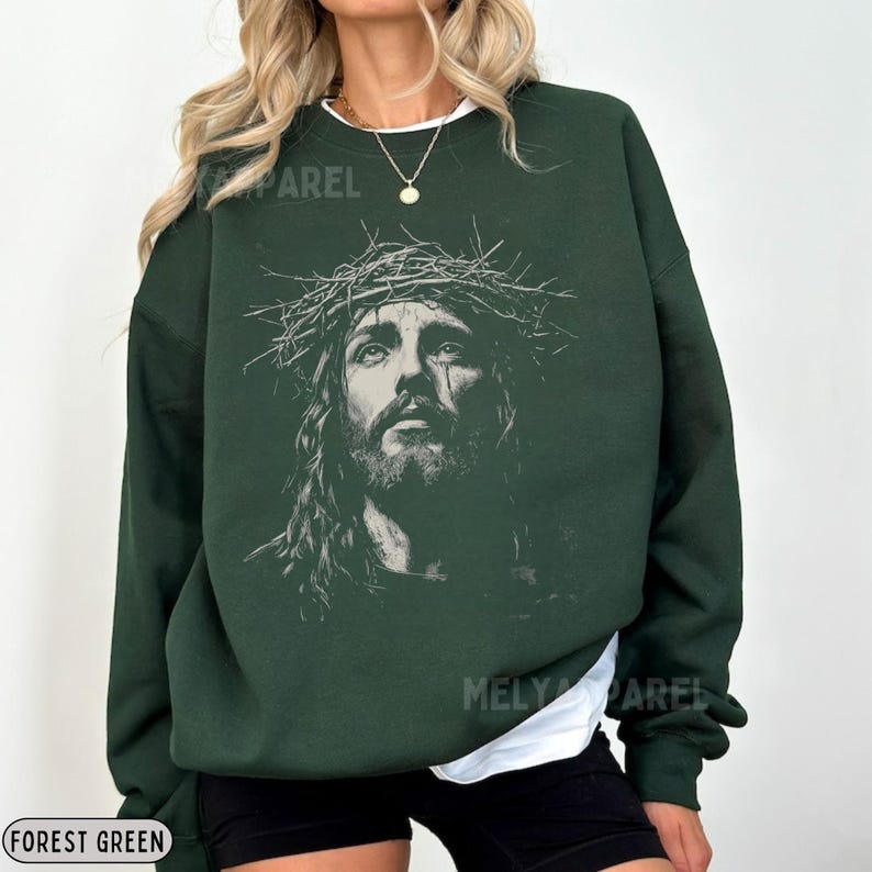 May include: Forest green sweatshirt featuring a detailed graphic of Jesus wearing a crown of thorns. The image is a grayscale portrait. The sweatshirt has a crew neck and long sleeves. The text "MELY APPAREL" is visible on the garment.