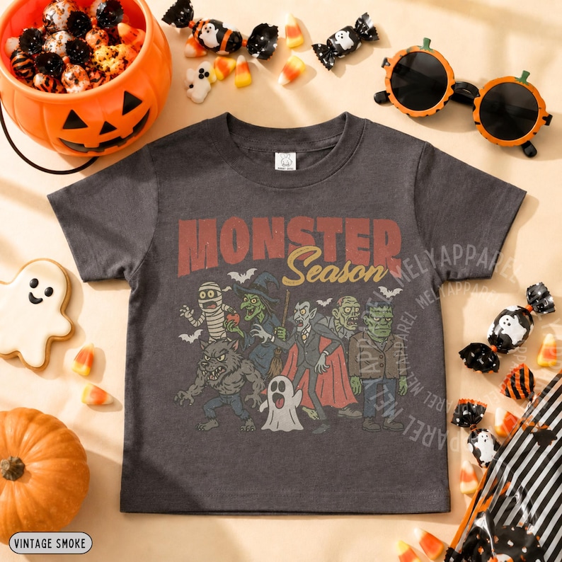 May include: A charcoal gray short-sleeve t-shirt featuring a graphic print with the text Monster Season and illustrations of a mummy, witch, vampire, werewolf, ghost, and Frankenstein's monster surrounded by small bats.