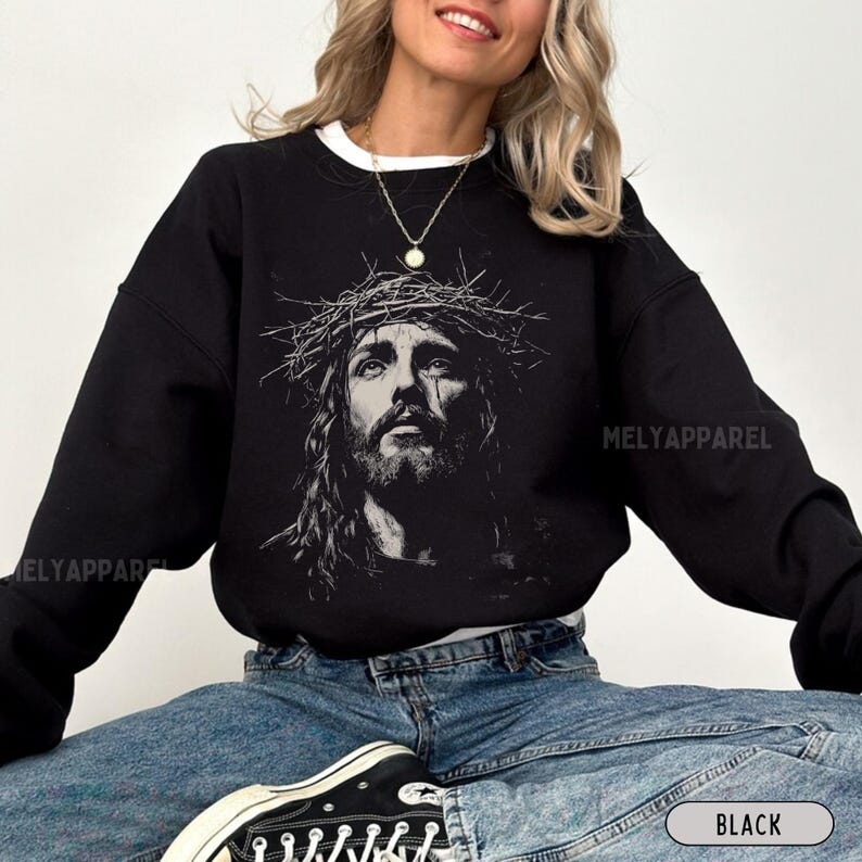 May include: Black sweatshirt featuring a grayscale image of Jesus wearing a crown of thorns. The sweatshirt has a crew neck and long sleeves. The text "MELYAPPAREL" is printed on the sleeve. The word "BLACK" is printed on the bottom right.