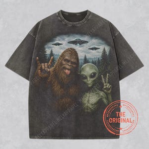 May include: A charcoal gray short-sleeve t-shirt featuring a graphic print of Bigfoot and an alien standing in a forest with three flying saucers above them. Bigfoot is making a rock-and-roll hand gesture, and the alien is making a peace sign.
