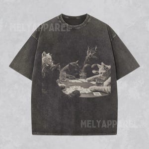 Puede incluir: Camiseta gris oscuro con un gr&aacute;fico en blanco y negro de gatos jugando al p&oacute;quer. La imagen muestra cinco gatos alrededor de una mesa con cartas y bebidas. La camiseta tiene un aspecto vintage y desgastado.