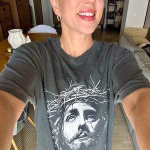 May include: Dark gray t-shirt featuring a white graphic of Jesus wearing a crown of thorns. The shirt has a crew neck and short sleeves. The image is a close-up of the face of Jesus.