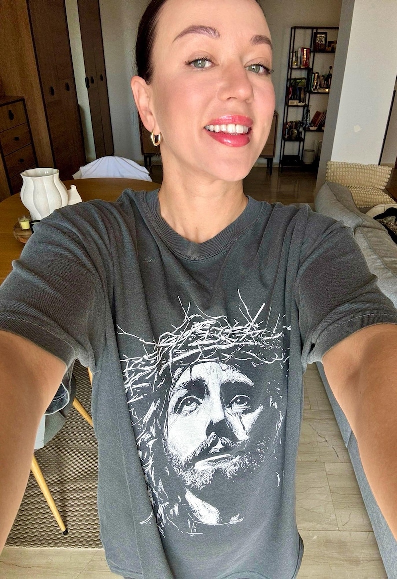 May include: Dark gray t-shirt featuring a white graphic of Jesus wearing a crown of thorns. The shirt has a crew neck and short sleeves. The image is a close-up of the face of Jesus.
