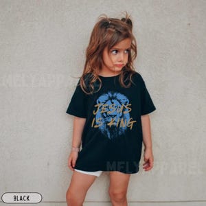 以下が含まれることがあります： 青いライオンのグラフィックと金色のテキスト「JESUS IS KING」が特徴の黒いTシャツ。若い人が着ています。カジュアルウェアです。