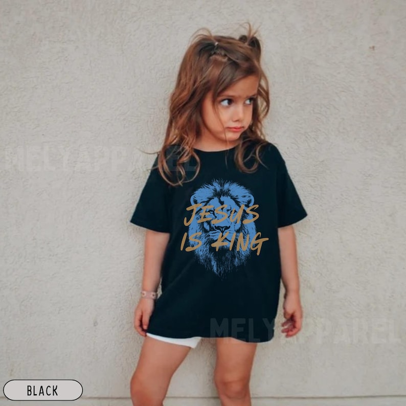 以下が含まれることがあります： 青いライオンのグラフィックと金色のテキスト「JESUS IS KING」が特徴の黒いTシャツ。若い人が着ています。カジュアルウェアです。