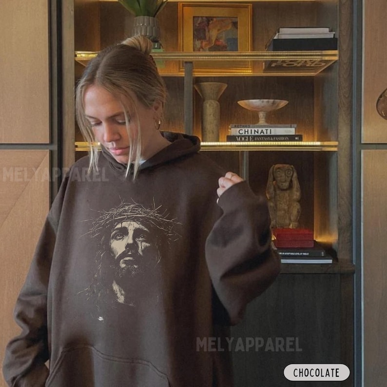 May include: Chocolate brown hoodie with a graphic of a man wearing a crown of thorns. The hoodie has a drawstring hood and a kangaroo pocket. The brand name "MELY APPAREL" is printed on the front. The word "CHOCOLATE" is in a white box.