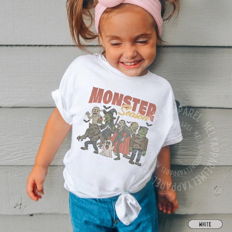 May include: A white short-sleeved t-shirt featuring a graphic print with the text MONSTER Season in brown and yellow. The illustration depicts a group of classic monsters including a mummy, witch, vampire, werewolf, ghost, and Frankenstein's monster.