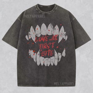 K&ouml;nnte beinhalten: Dunkelgraues T-Shirt mit einem verwitterten Aufdruck eines Mundes mit scharfen Z&auml;hnen und den Worten "LOVE AT FIRST BITE" in Rot. Das Shirt hat einen Vintage-Look und der Markenname "MELYAPPAREL" ist oben aufgedruckt.